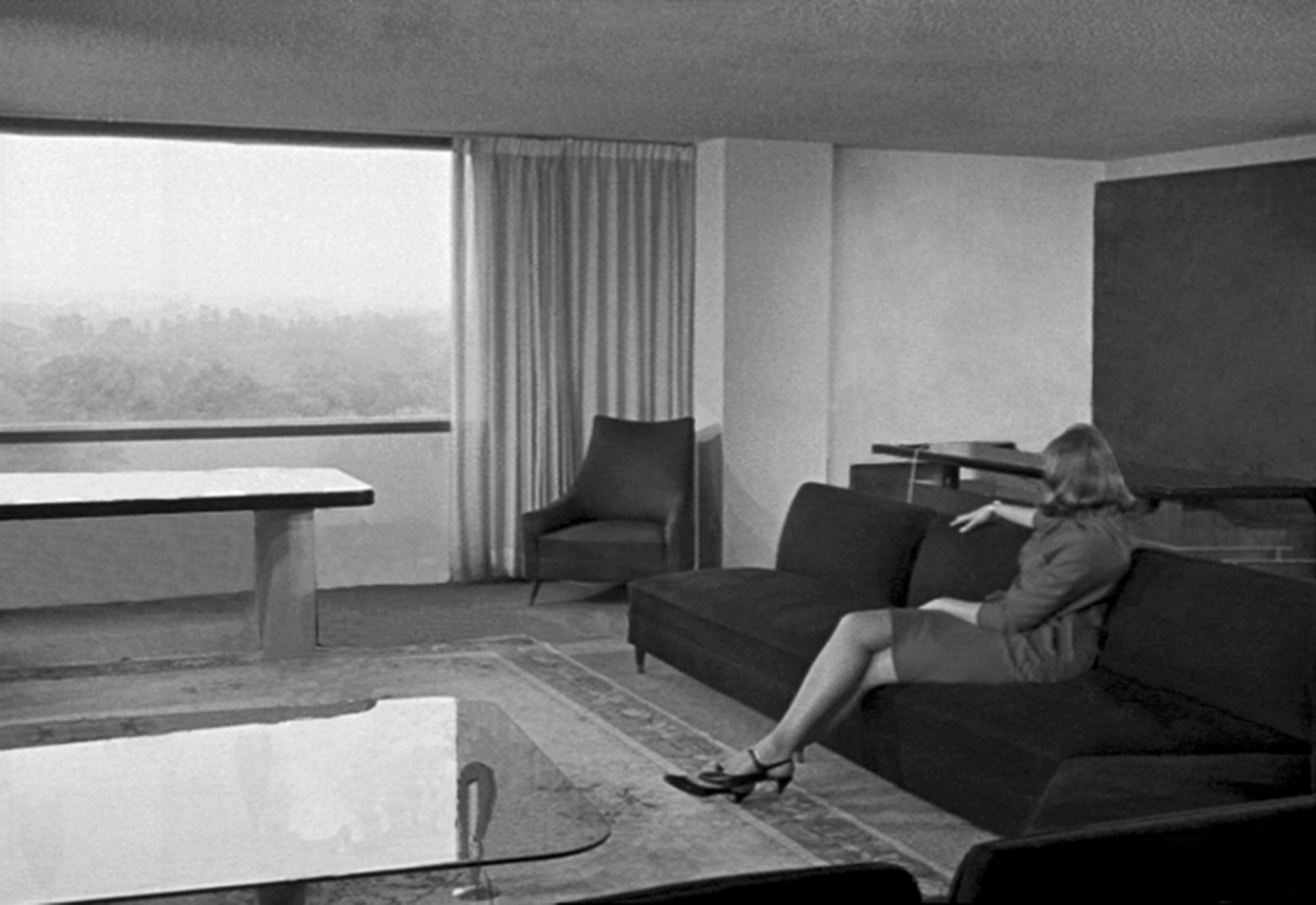 A film still shows a suit-clad woman staring out of the picture window of a modernist home.