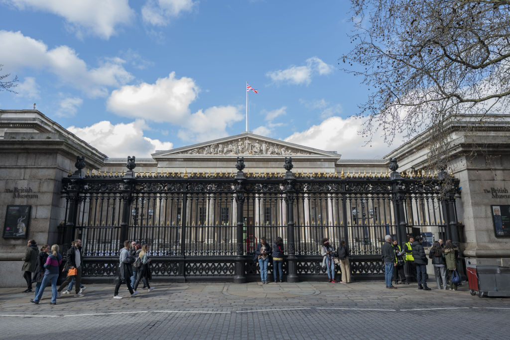 The front of the British museum behind a tall iron gate with people walking in front of it and peering inside.