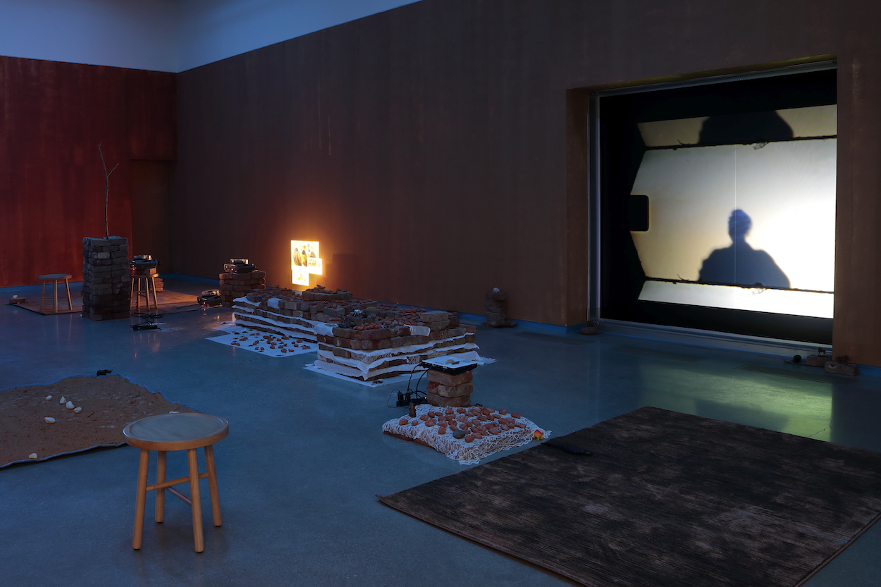 A darkened gallery contains a number of sculptures and objects, including a soil-covered blanket, a stool, and several sculptures low to the ground, as well as a projected video and projected slide images.