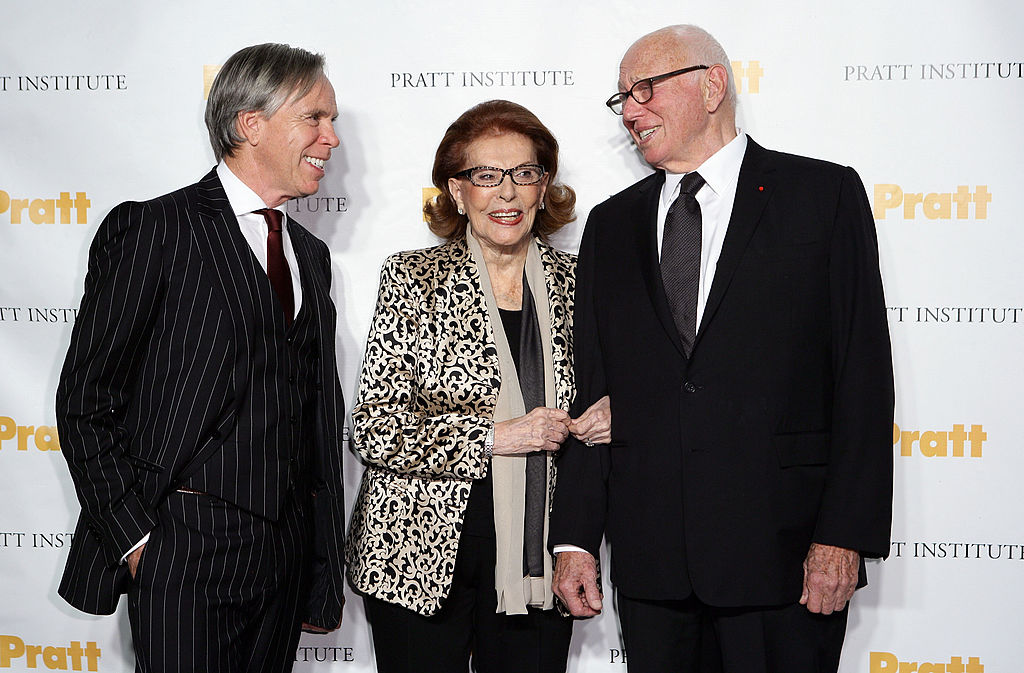 Two men in suits stand on either side of a woman in a patterned jacket. A backdrop behind them repatedly reads "Pratt Institute" and "Pratt."