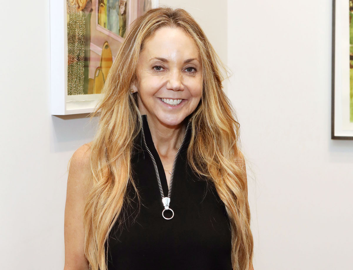 Portrait of a smiling white woman in a black top with a long zipper running down its middle. Behind her are abstract paintings.
