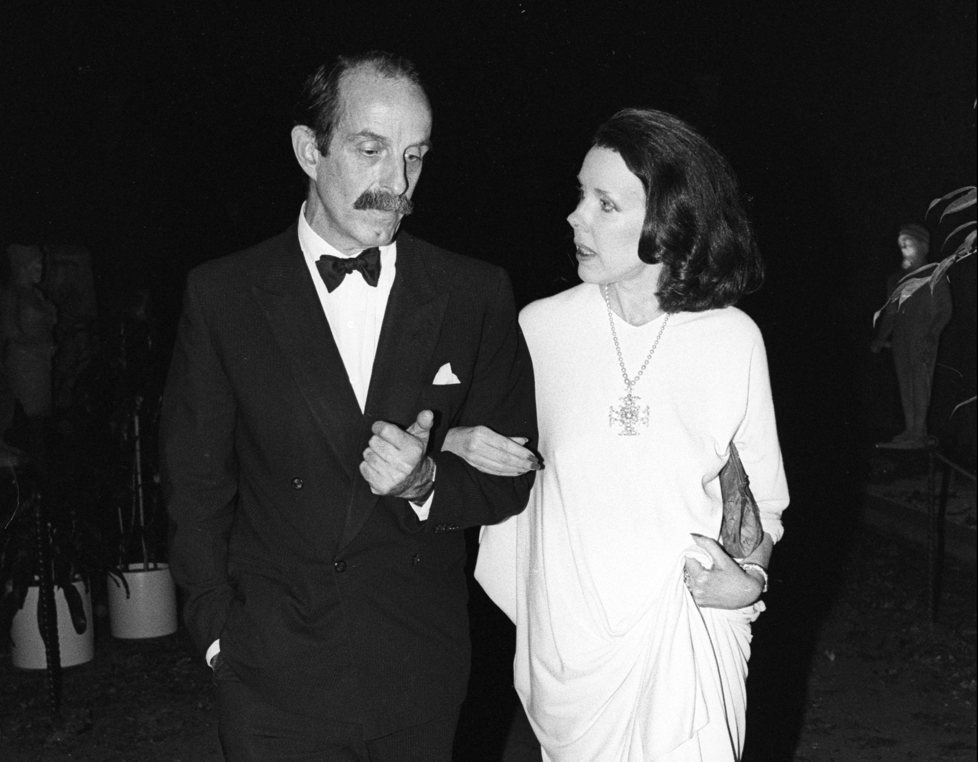 In a black-and-white photograph, a man in a tuxedo and bow tie and a woman in a long white dress walk arm in arm.