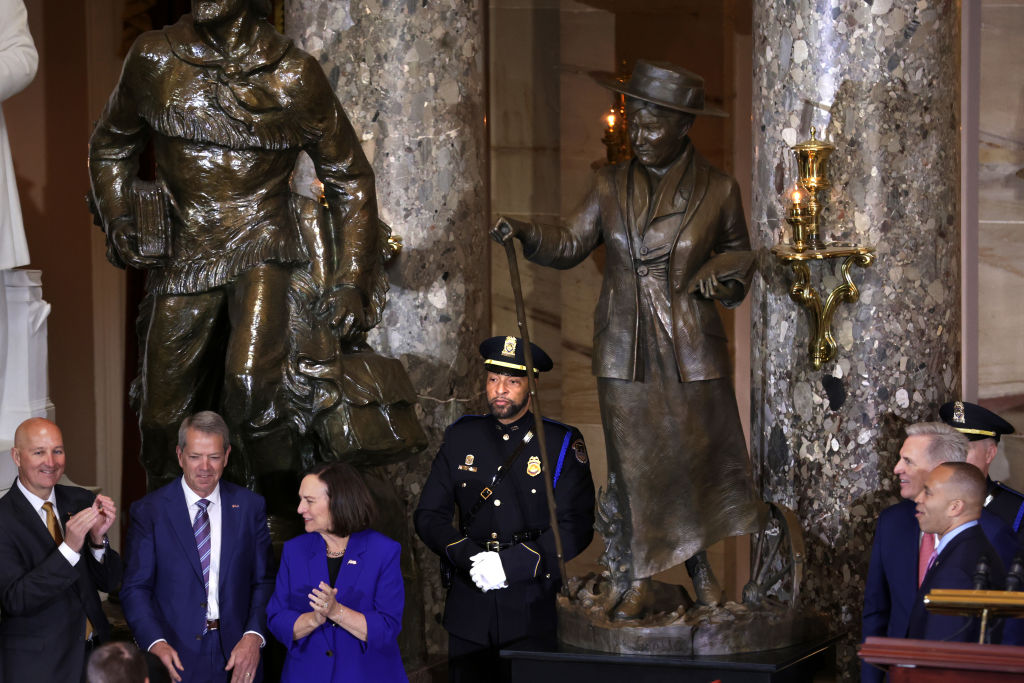 Five people in suits and two police officers in uniform stand in a grand hall. Tall statues loom over them.