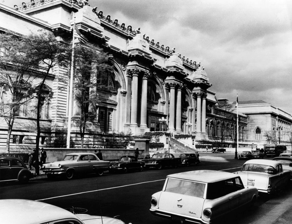In a black-and-white photo, a cars are parked in front of a grand Neoclassical building.