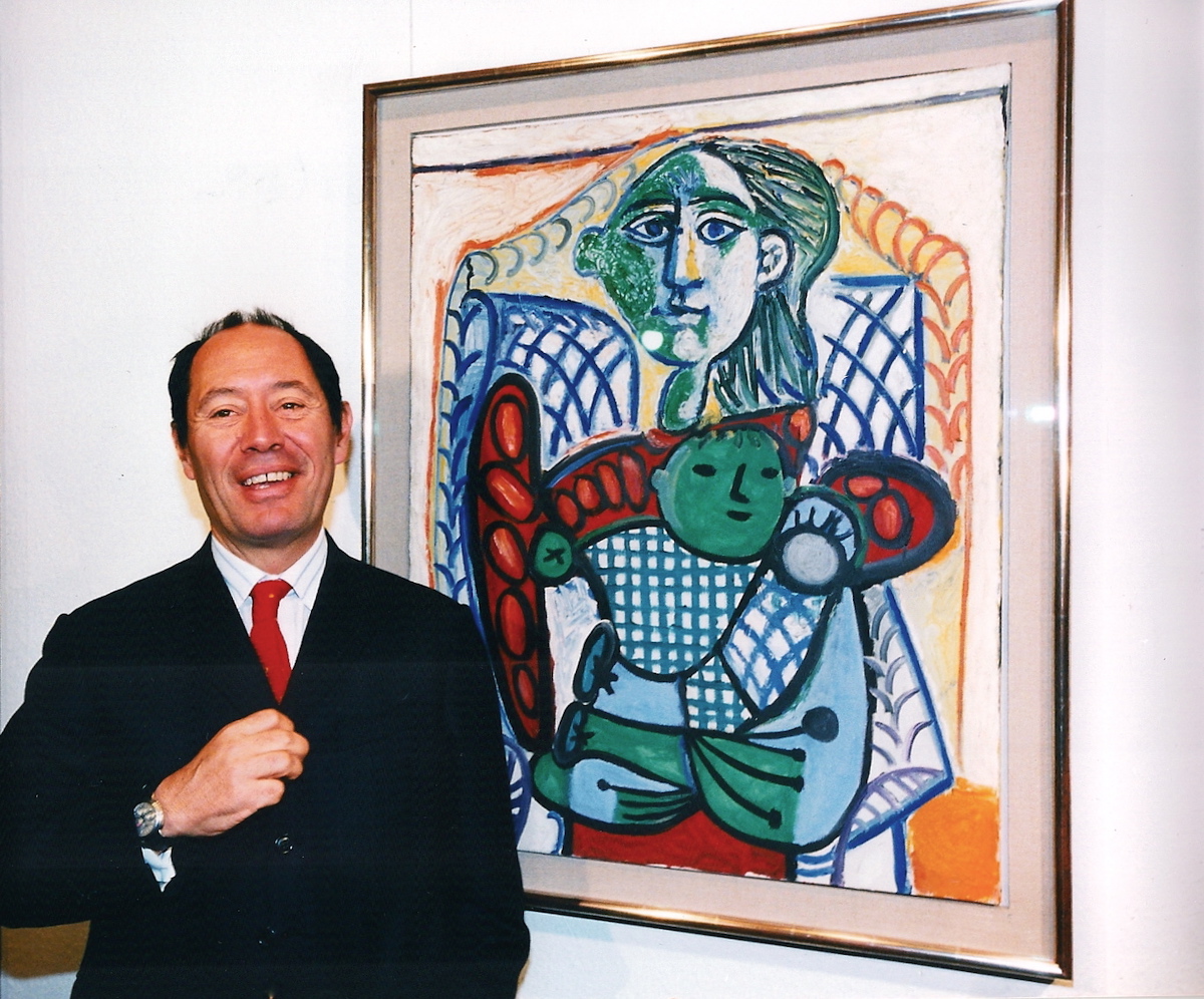 A smiling man in a suit beside an abstracted painting of a woman with a baby.