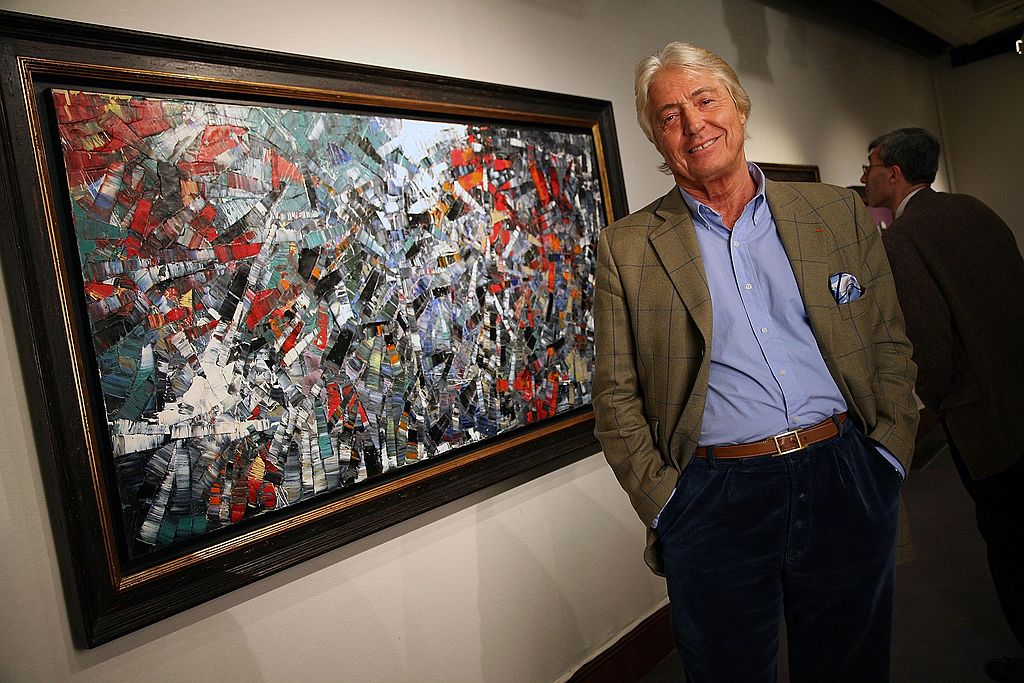 An older gentleman in an olive blazer, blue shirt, and dark pants poses with his hands in the pockets of his pants next to a collage-style painting.