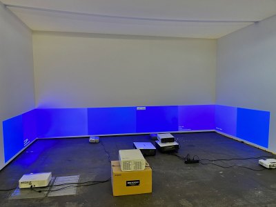 Installation view of a several projectors on the floor which all show a blue calibration screen. Each blue is a slightly different hue from the others.
