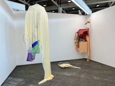 Two textile-based sculptures hang from the ceiling of an art fair booth, with a glass sculpture filled with white soap rests on the floor.