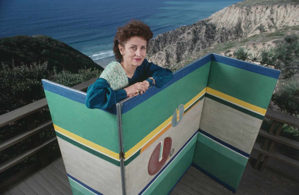 A white woman with brown hair behind a green folding screen against a backdrop of a seascape.
