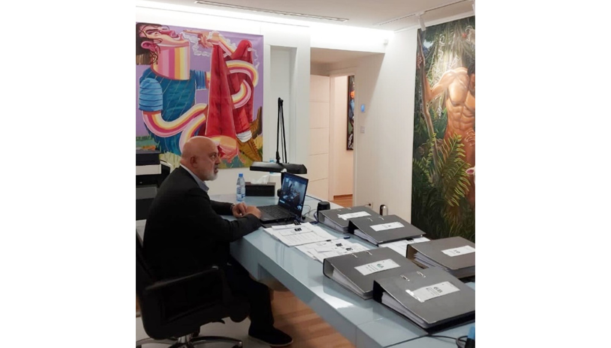 Art collector Nazem Ahmad sits in an office chair in front of a laptop, papers, and several thick black binders on a long off-white desk. Two large paintings have been hung on the walls to his left and in front of him. Nazem is staring forward and wearing a suit with casual shoes.
