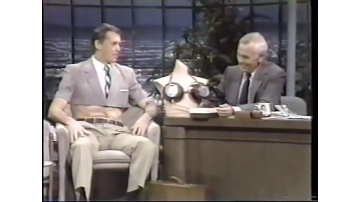 A muscsular man appears on the Tonight show wearing a suit, but the top half is cut off like a crop top, exposing his abs. Johnny Carson is sitting next to him at a desk, where a mannequin wearing a bra made out of pans rests.