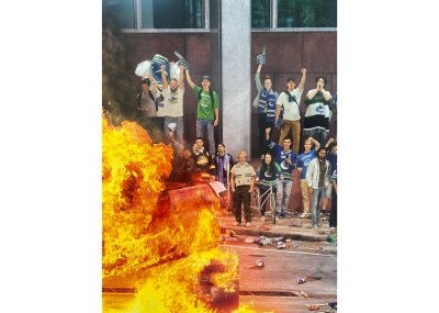 A digitally altered photograph showing various people who are dressed for a sporting event looking at a car on fire.