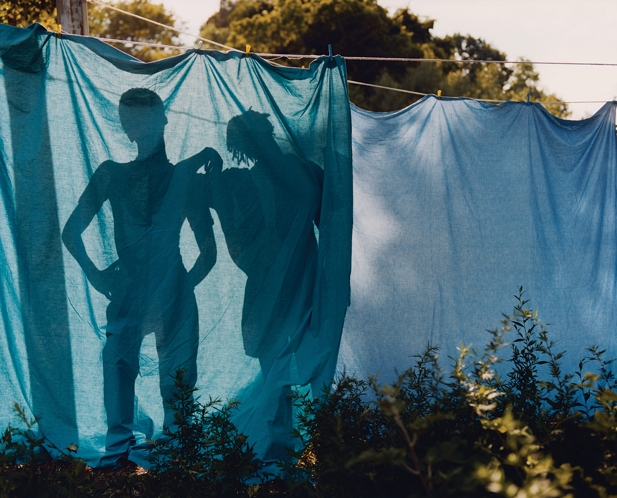 Two shadowy figures that look like young boys are visible through two blue sheets hanging on a laundry line.