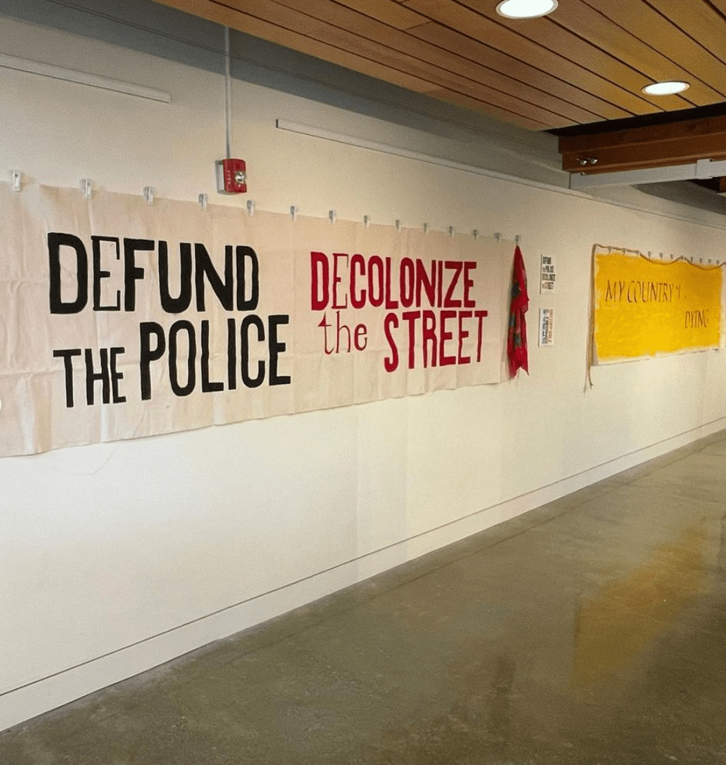 A banner reading DEFUND THE POLICE / DECOLONIZE THE STREET pinned to a gallery wall.